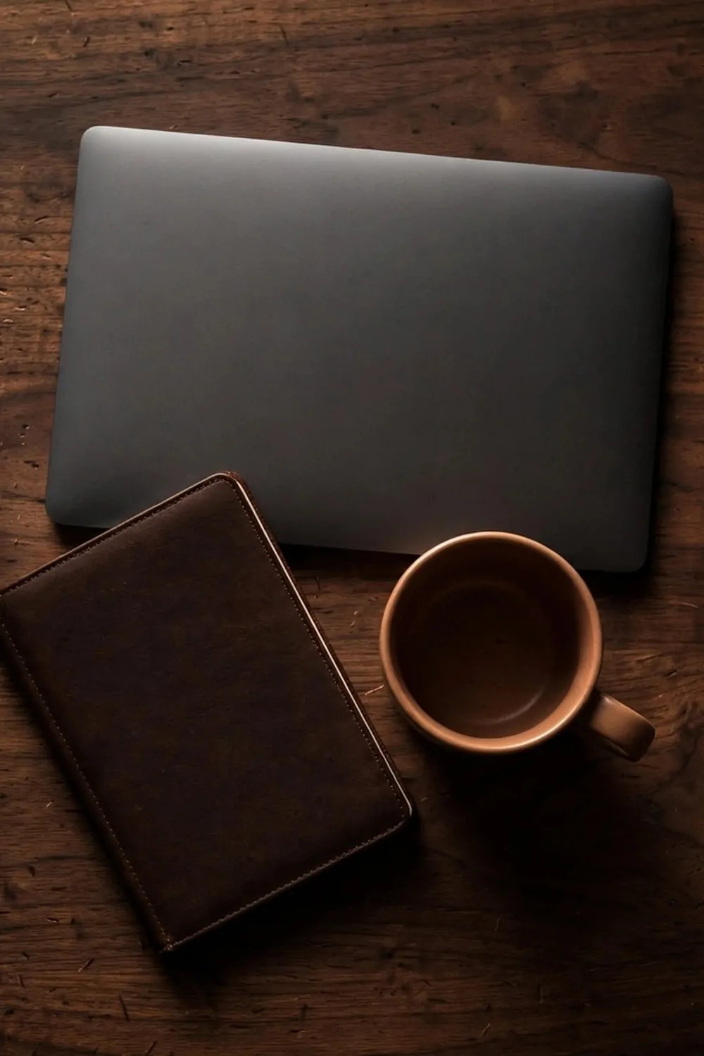 17 Dark Cozy Office Ideas With Moody, Rich Textures 3 A dark walnut wooden desk top with a closed laptop, notebook and mug, warm moody light