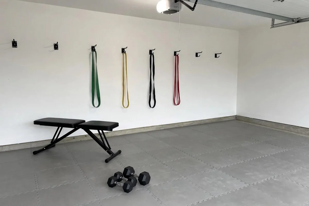 Minimal starter garage gym with a folding bench, dumbbells and bands on foam tiles