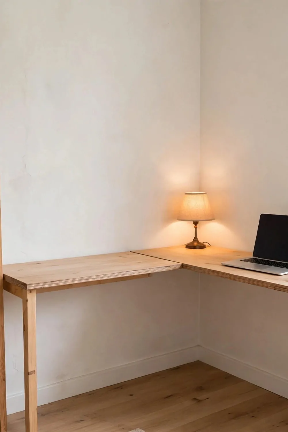 20 Extra Bedroom Office Ideas to Use Every Spare Inch 4 A corner desk made of two slim wood boards meeting at a right angle in a spare bedroom corner, laptop on the desk, small warm lamp at the far end, tall shelf on one side with books, bright room