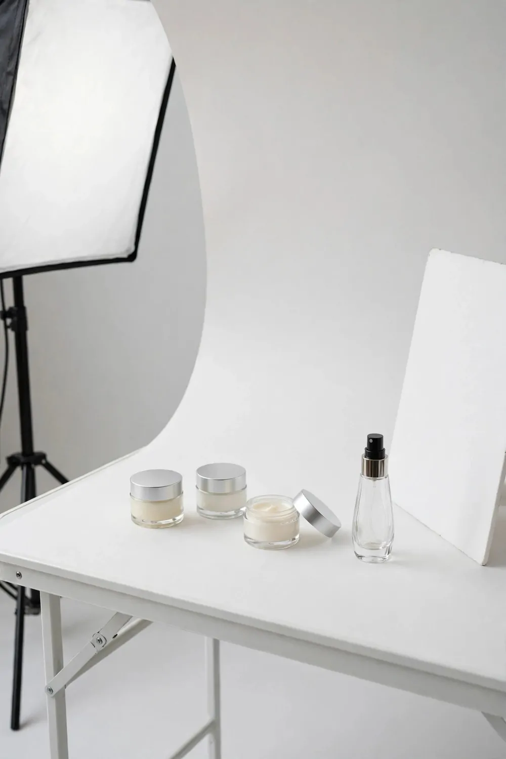 A tabletop product photography station with cosmetic jars on a white table, seamless paper background, softbox light and reflector card