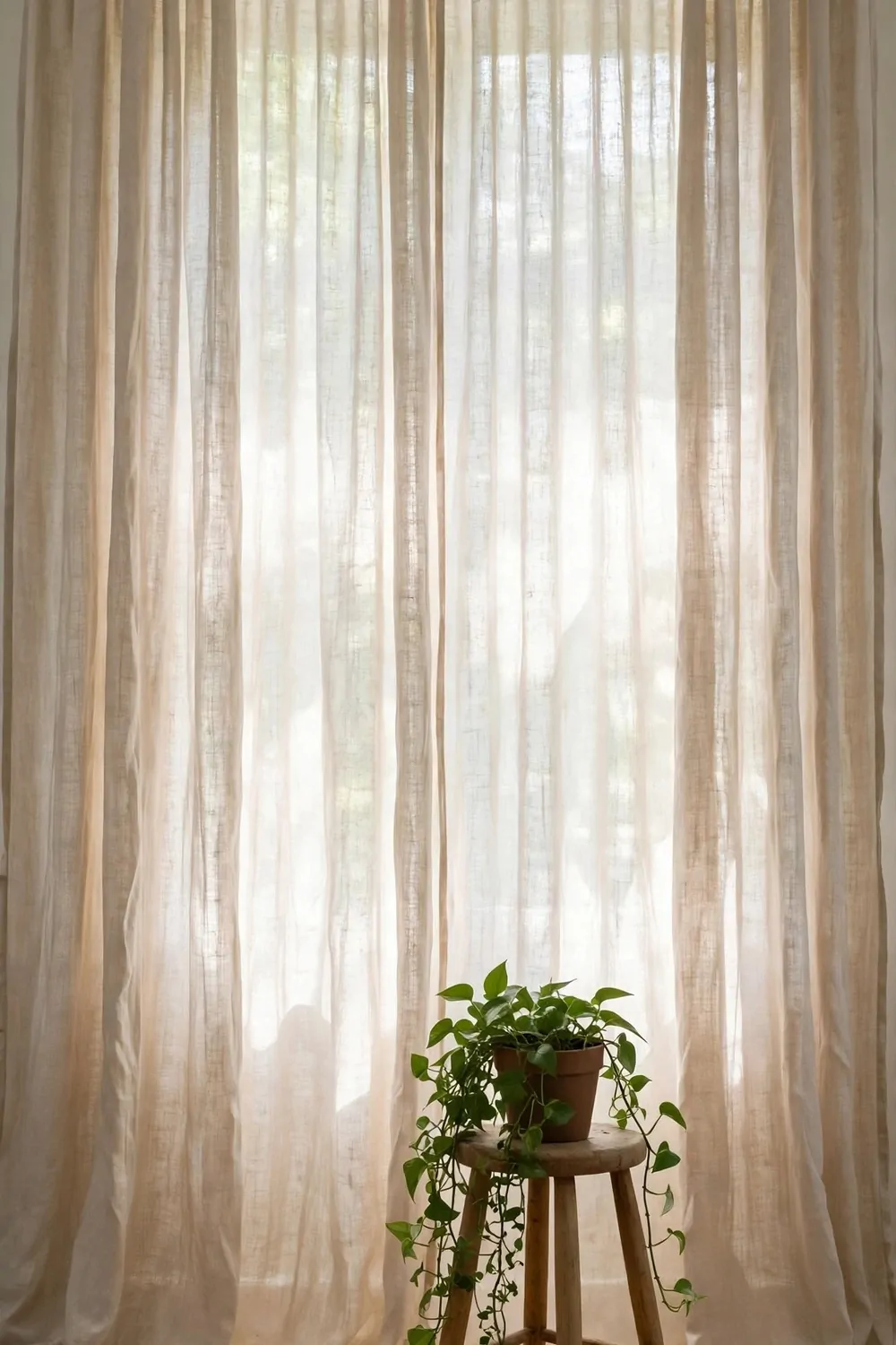 A floor-to-ceiling natural linen curtain backdrop with a wooden stool and trailing plant in front, soft window light filtering through