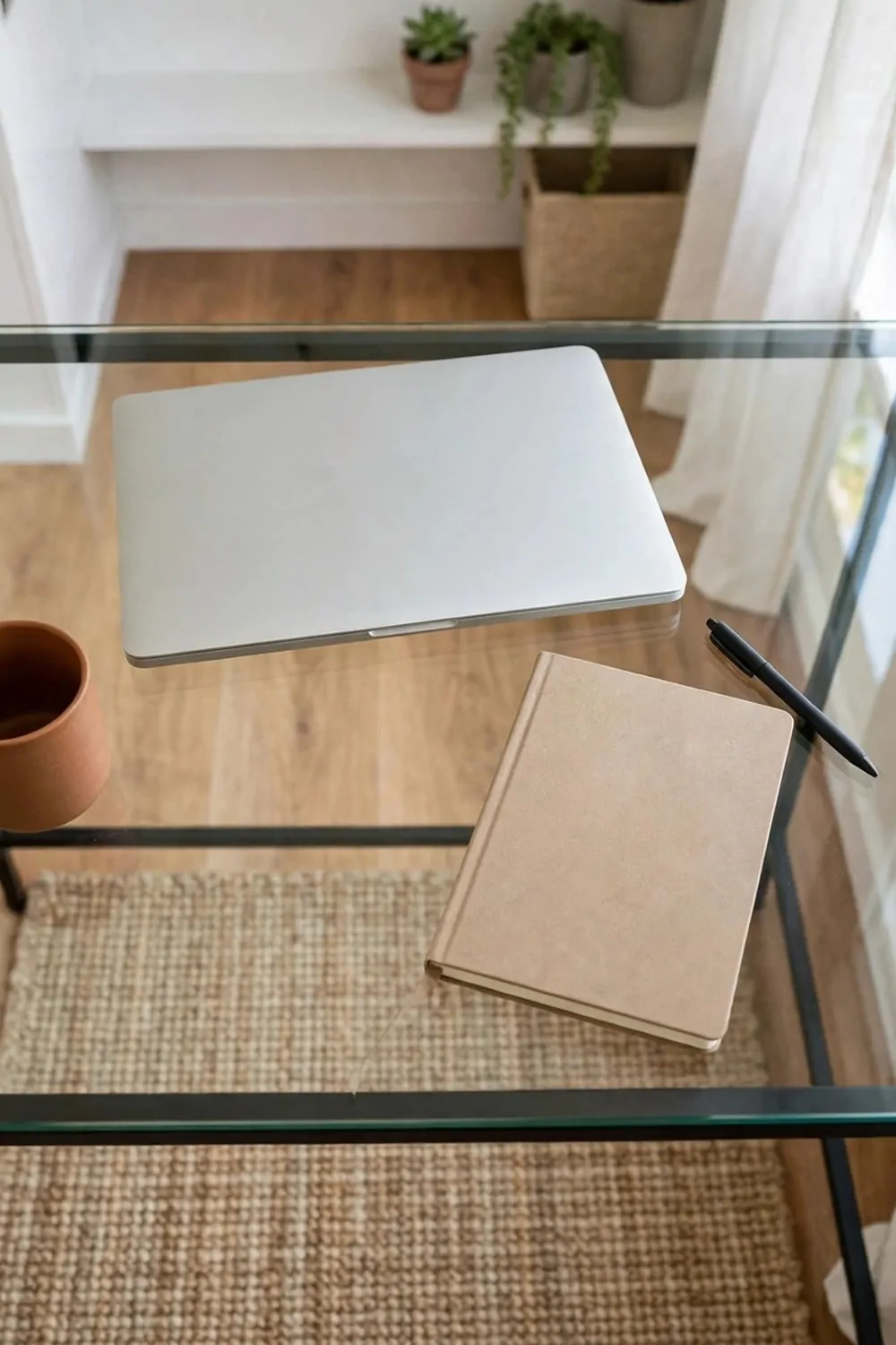 A glass-top desk in a tiny home office, laptop and notebook on the clear glass, floor visible through the desk, bright room